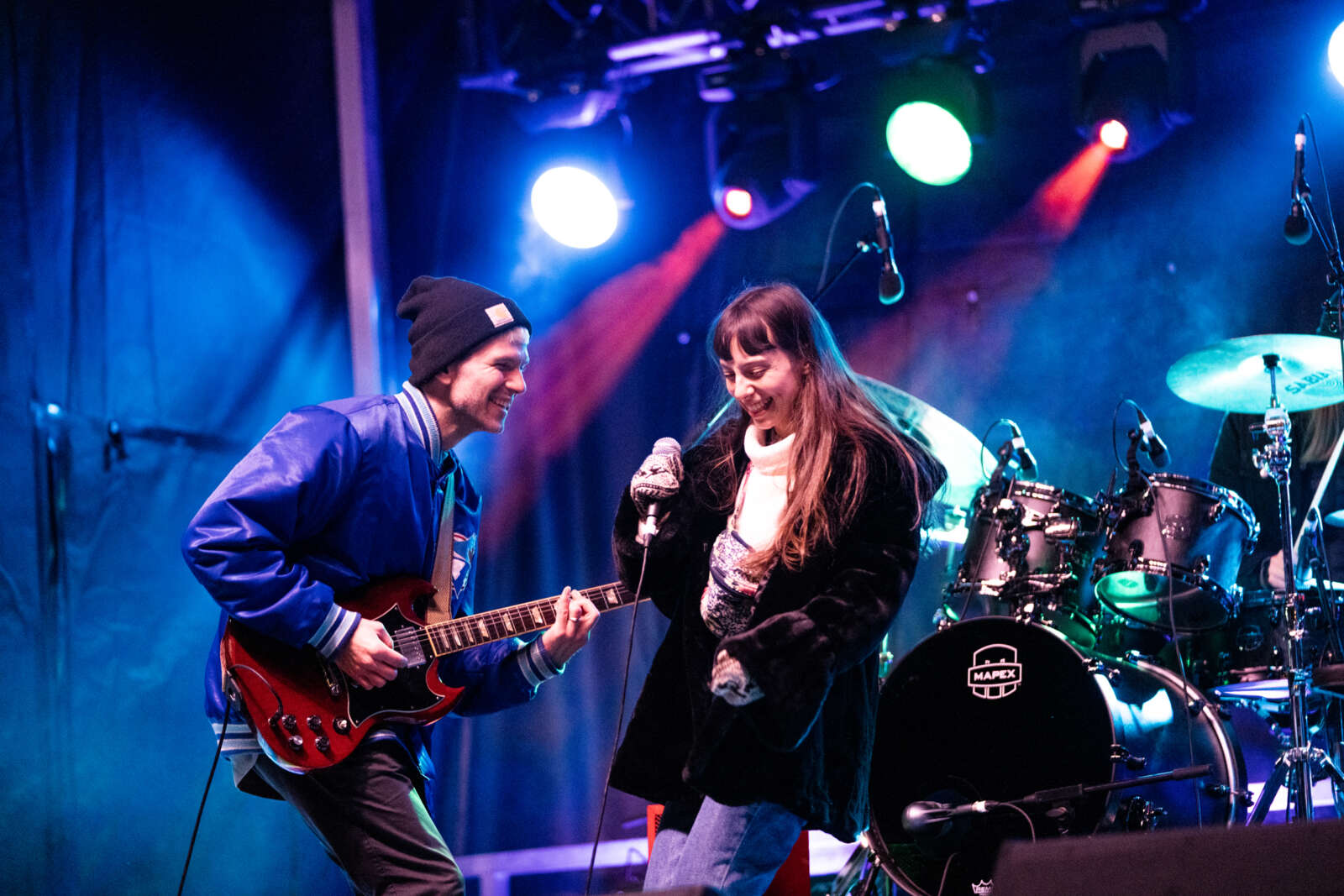 Man playing guitar and woman singing on stage outside with drumb set in background